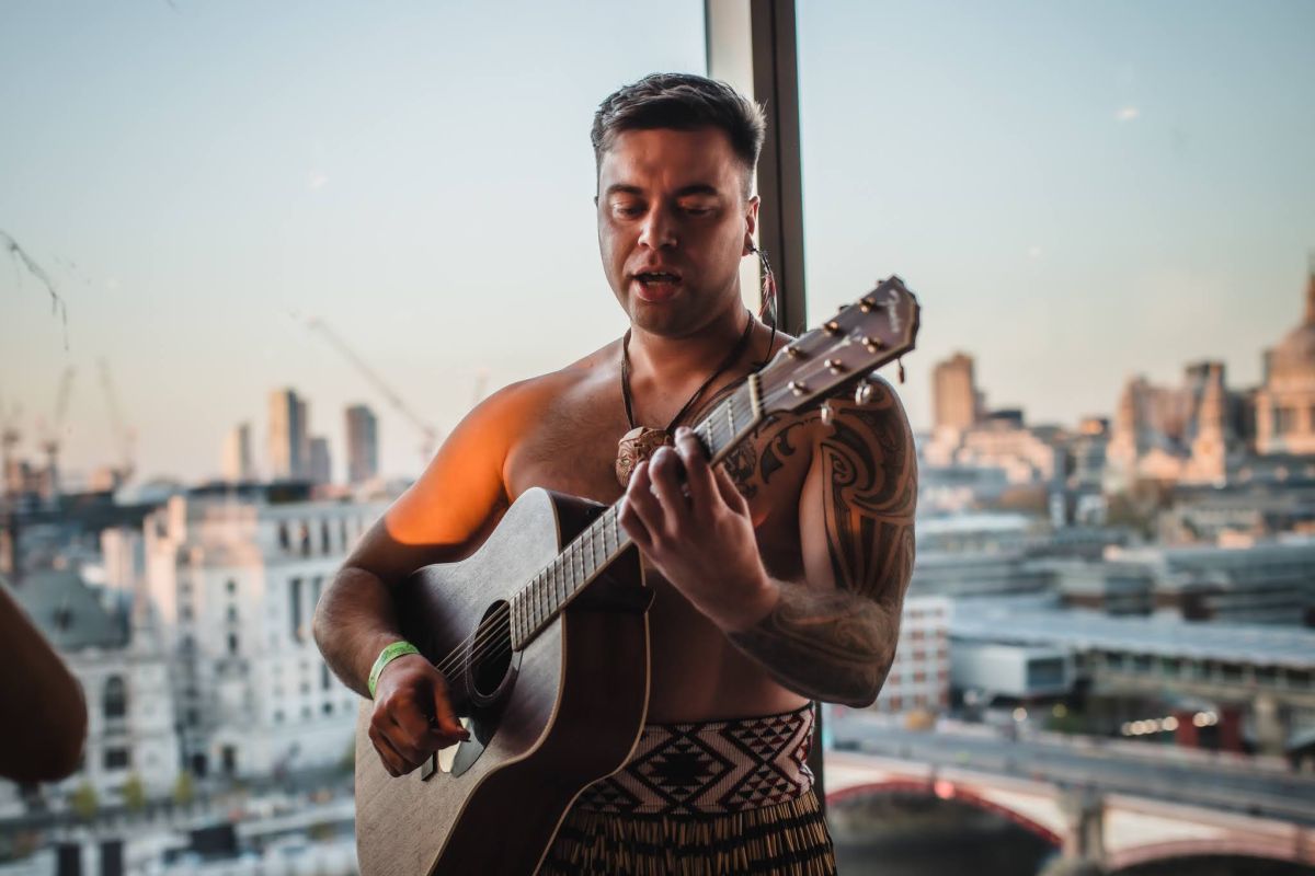 A Haka International performer singing waiata with a guitar, wearing a piupiu and pounamu , with the London skyline and the River Thames behind him